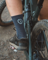 Side view of charcoal blue Trail Crew Socks on model, mid-calf ribbed knit with logo shown on a muddy mountain bike shoe