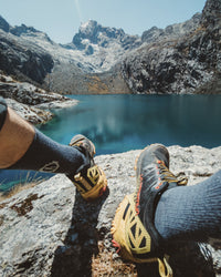 Hiker’s legs wearing charcoal blue crew socks and trail shoes resting on a rock by a mountain lake
