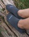 Angled top view of Trail Ankle Socks showcasing low-cut ankle fit and white PAKA logo on charcoal alpaca-blend fabric against a weathered log