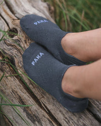 Angled top view of Trail Ankle Socks showcasing low-cut ankle fit and white PAKA logo on charcoal alpaca-blend fabric against a weathered log