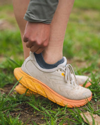Side view of Trail Ankle Socks in charcoal blue on a foot showing low-cut ankle fit and heel tab with grassy trail background