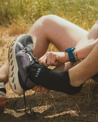 Side view of black Trail Ankle Socks on a runner’s foot showing PAKA logo and low-cut ankle fit on grassy trail