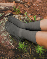 Side view of charcoal Trail Crew Socks on model showing ribbed knit texture, calf-length fit and PAKA logo on toes with rocky trail background
