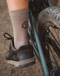 Side view of Trail Crew Socks in walnut on a model’s ankle showing ribbed knit and logo with mountain bike and tire background