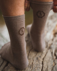Rear view of Trail Crew socks in walnut showing mid-calf fit and PAKA logo on cuff, worn standing on a weathered log