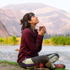 Woman sitting cross legged in front of river with mountains in background holding a cup of coffee