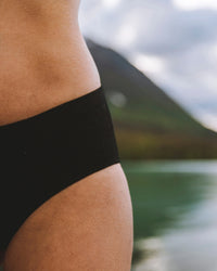 Close-up of Women’s Essential Mid-Rise Brief in black showing mid-rise cut and smooth alpaca fabric on model with lake behind