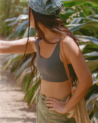 Side view of Women’s Essential Scoop Bralette in Forage on model, fitted, showing scoop neckline, wide straps, and breathable alpaca fabric against a sunlit leafy background