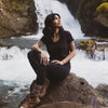 A woman sitting in the rocks in front of a waterfall. She's wearing a black alpaca v-neck tee