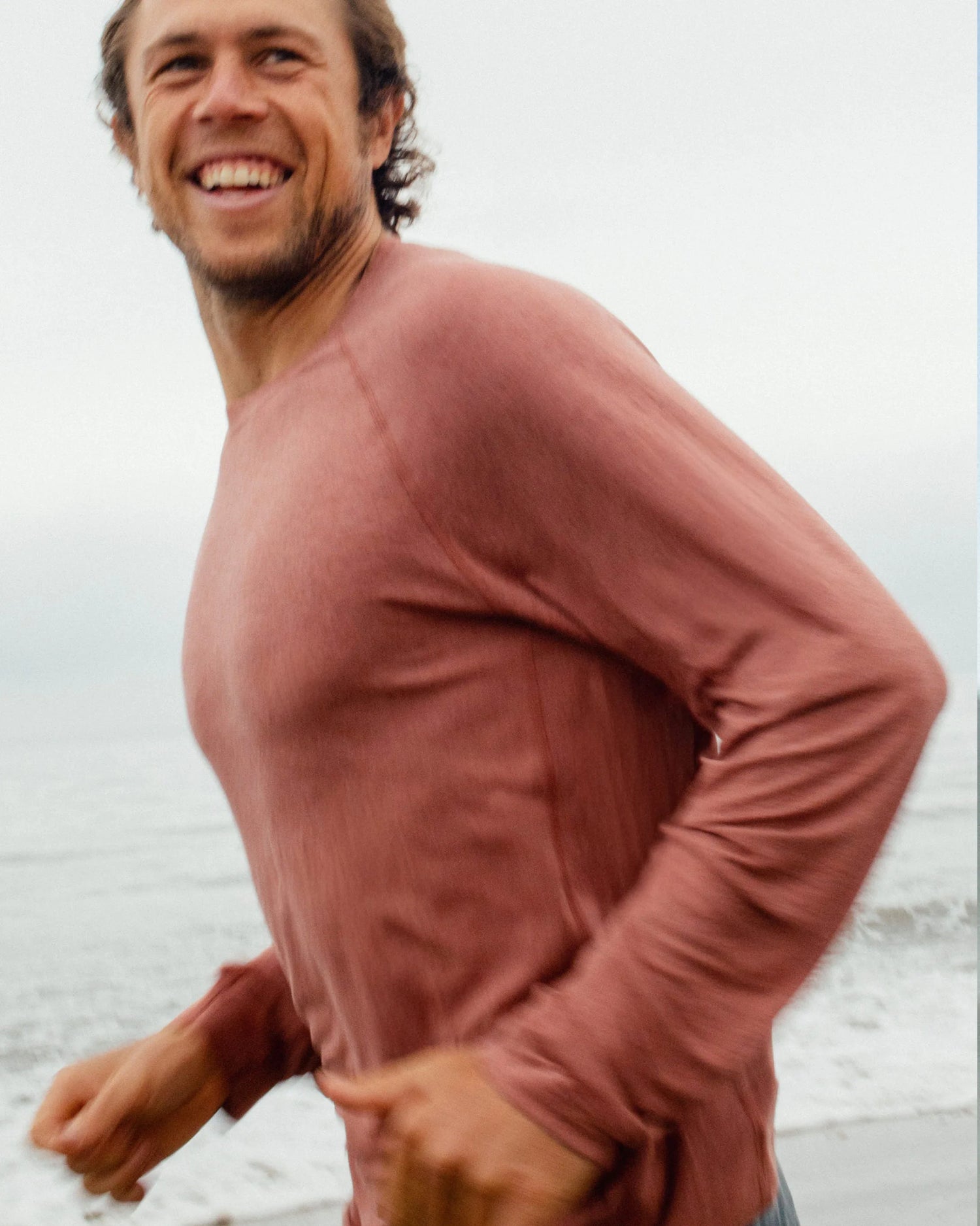 Man wearing a pink long-sleeve shirt standing on a beach with a light blue sky.