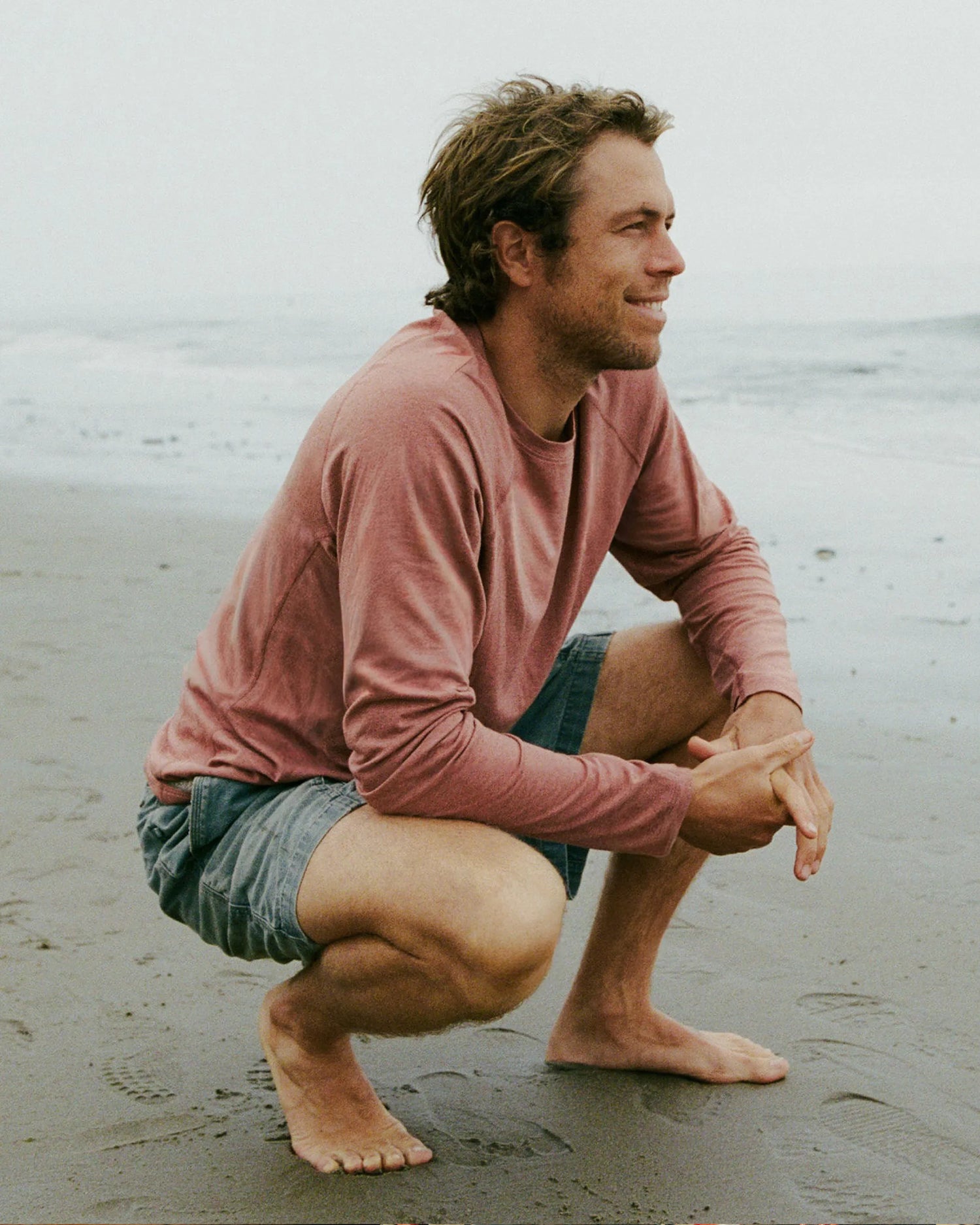 Man squatting on a beach wearing a pink long-sleeve shirt and denim shorts.