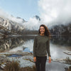 A smiling woman wearing a green alpaca Crewneck in front of a lake and mountains. 