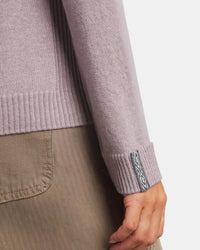 Close-up of a person wearing a light purple sweater with a brand logo on the sleeve, against a neutral background.