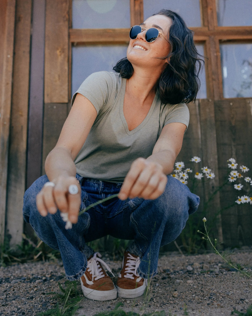 Woman squatting outdoors with sunglasses, wearing PAKA Women's Essential Tee in Willow color and blue jeans, surrounded by flowers.