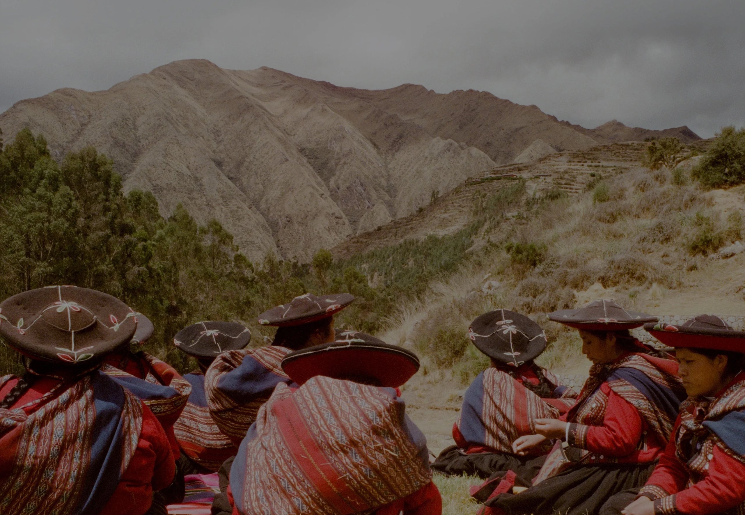 Group of Peruvian women in embroidered hats and shawls sitting on a hillside with terraced Andean mountains behind them