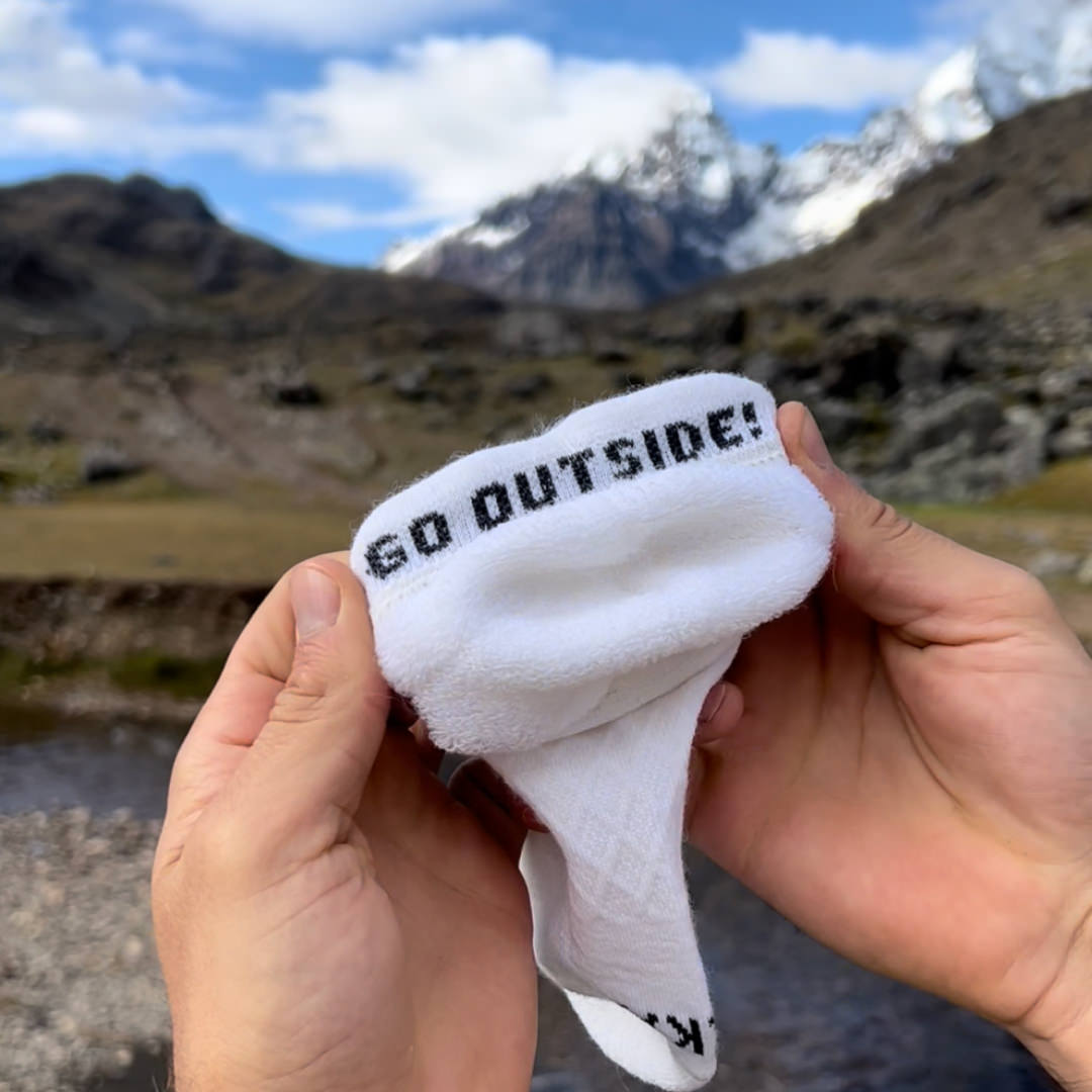A white alpaca sock in front of a mountain. It says: "go outside!"