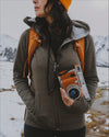 A woman wearing our green Women's Full Zip, holding a camera in a snowy landscape