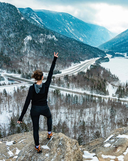 Woman Hiker in Lavender / Light Purple Alpaca Crew Socks Showing Peace Sign on Rocky Overlook of Snowy Valley and Roads – PAKA Apparel